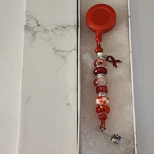Può includere: Un porta badge retrattile rosso con un ciondolo a forma di perline con perline rosse, bianche e trasparenti. Il ciondolo include anche un piccolo nastro rosso.
