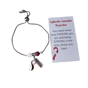 Può includere: Un braccialetto regolabile argentato con una perla bianca e viola, un ciondolo a nastro e un piccolo ciondolo in metallo con la scritta "WARRIOR". Una carta accanto al braccialetto recita "Aplastic Anemia Warrior".