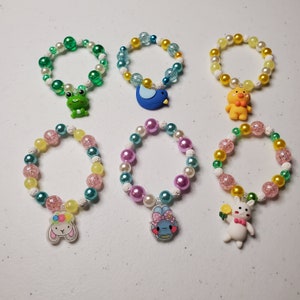 Puede incluir: Seis pulseras de cuentas con abalorios. Las pulseras presentan una mezcla de cuentas coloridas, incluyendo verde, blanco, rosa y amarillo. Los abalorios incluyen una rana, un pájaro, un pato y conejos. Las pulseras están diseñadas para niños.