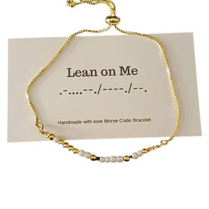 May include: Gold chain bracelet with white beads spelling out "Lean on Me" in Morse code. The bracelet is adjustable with a sliding knot.