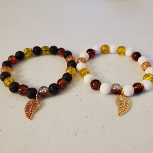 Puede incluir: Dos pulseras de cuentas con dijes de hoja de oro. Una pulsera tiene cuentas negras, ámbar y amarillas. La otra pulsera tiene cuentas blancas, ámbar y amarillas.