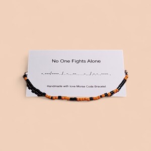 Könnte beinhalten: Ein handgefertigtes Morsecode-Armband mit schwarzen und orangefarbenen Perlen wird vor einer weißen Karte gezeigt. Die Karte trägt die Aufschrift "No One Fights Alone" mit Morsezeichen und "Handmade with love Morse Code Bracelet".