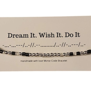 Puede incluir: Pulsera de cuentas negras y blancas con el mensaje "Dream It. Wish It. Do It." en código Morse. La pulsera está hecha a mano con amor.