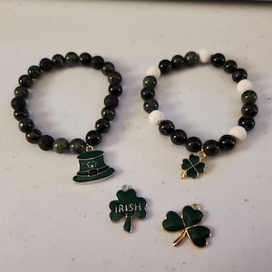 May include: Two green and black beaded bracelets with charms. One bracelet has a white lava bead and a shamrock charm. The other bracelet has a green leprechaun hat charm. There are two additional shamrock charms, one with the word "IRISH" on it.
