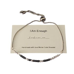 Op de afbeelding: Een zilverkleurige Morsecode-armband met zwarte en zilveren kralen. De armband wordt getoond voor een kaart met de tekst "I Am Enough" en "Handmade with love Morse Code Bracelet".