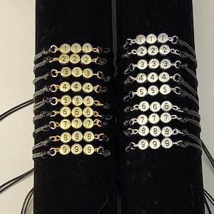 May include: Two rows of black string bracelets with gold and silver beads. Each bracelet has three beads with a number from 1 to 9 printed on each bead. The bracelets are displayed on a black velvet background.