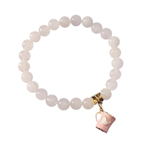 Puede incluir: Un brazalete de cuentas blancas con un dije dorado en forma de taza de café rosa con un corazón blanco.