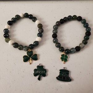 May include: Two green and black beaded bracelets with shamrock charms. One bracelet has white beads and the other has black beads. The charms are gold with green enamel.