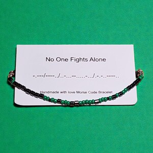 Könnte beinhalten: Ein handgefertigtes Morsecode-Armband mit schwarzen und grünen Perlen wird auf einer weißen Karte gezeigt. Auf der Karte steht "No One Fights Alone" mit Morsezeichen. Das Armband befindet sich auf grünem Hintergrund.
