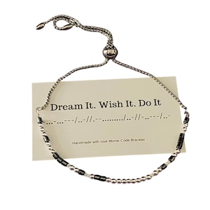 Puede incluir: Un brazalete de cuentas plateadas y negras con un cierre deslizante. El brazalete tiene una tarjeta adjunta que dice "Dream It. Wish It. Do It." y "Handmade with love Morse Code Bracelet".