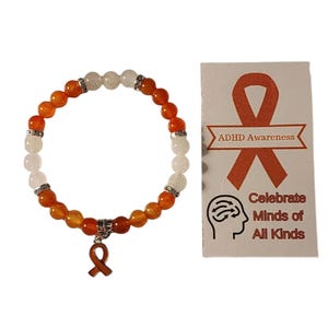 Peut inclure: Bracelet en perles orange et blanches avec un petit charme en ruban orange. Le bracelet est accompagné d'une carte qui dit "Sensibilisation au TDAH" et "Célébrer les esprits de toutes sortes".