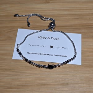 May include: A silver-toned adjustable bracelet featuring a series of small metallic silver and black cylindrical beads, centred with a black cat-shaped charm. The bracelet is displayed on a white card labelled Kirby & Dude with Morse code.