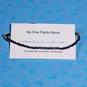 Könnte beinhalten: Ein handgefertigtes Morsecode-Armband mit schwarzen und blauen Perlen, präsentiert auf einer Karte mit der Aufschrift "No One Fights Alone". Das Armband besteht aus kleinen Perlen, vor blauem Hintergrund.
