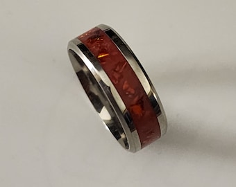 Puede incluir: Un anillo plateado con una banda ancha. El centro del anillo presenta una incrustación rojo-anaranjada vibrante con una apariencia texturizada. El diseño del anillo es simple y moderno, adecuado para el uso diario.