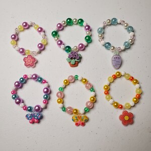 Op de afbeelding: Zes kleurrijke kralenarmbanden met bedels. De bedels zijn een bloem, een plant in een pot, een wortel, een vlinder en een bloem. De armbanden zijn gemaakt van witte, roze, groene, blauwe en gele kralen.