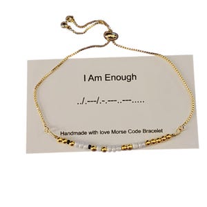 Op de afbeelding: Een gouden kettingarmband met witte en zwarte kralen die "Ik ben genoeg" in morsecode spellen. De armband ligt op een witte kaart met dezelfde boodschap en de woorden "Handgemaakt met liefde Morsecode armband".