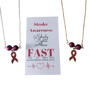 May include: Two necklaces with a Stroke Awareness theme. Each necklace has a chain, two dark pink beads, and a red ribbon charm. A card with the text "Stroke Awareness" and "FAST" is also visible.