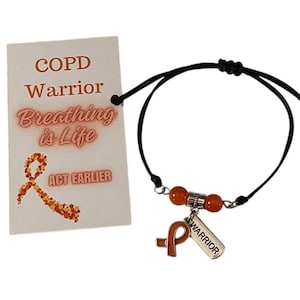 Puede incluir: Pulsera con cordón negro y cuentas, adornada con un dije de cinta naranja. La tarjeta adjunta dice "COPD Warrior Breathing is Life Act Earlier".
