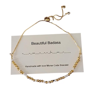 May include: Gold and white beaded bracelet with an adjustable sliding clasp. The bracelet has a card attached that says "Beautiful Badass" and "Handmade with love Morse Code Bracelet".