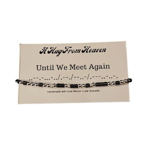 May include: A white card with black text that reads "A Hug From Heaven" and "Until We Meet Again". Below the text is a black and silver beaded bracelet with the message in Morse code. The card says "Handmade with love Morse Code Bracelet".
