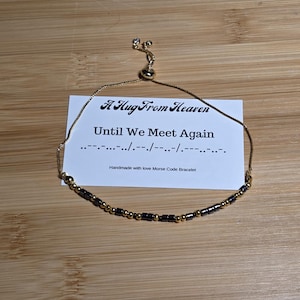 May include: A gold-toned Morse code bracelet with black and gold beads, displayed on a wooden surface. The bracelet is positioned near a card inscribed with "A Hug From Heaven" and "Until We Meet Again". The card also says "Handmade with love Morse Code Bracelet".