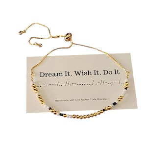 Puede incluir: Pulsera de cuentas doradas con cuentas blancas y negras. La pulsera tiene un cierre deslizante y una tarjeta que dice "Dream It. Wish It. Do It." en texto negro. La tarjeta también incluye el código Morse para la frase y dice "Handmade with love Morse Code Bracelet".