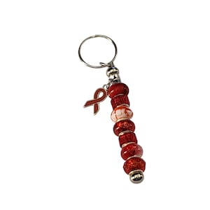 Può includere: Un portachiavi con perline rosse e bianche, un anello argentato e un ciondolo a forma di nastro rosso.