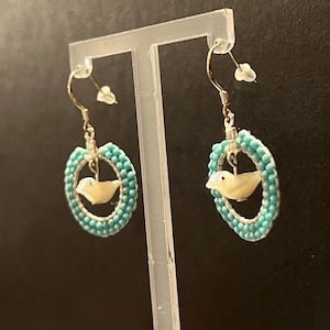 May include: A pair of dangle earrings featuring a small, off-white bird pendant within a turquoise beaded oval. The earrings have gold-colored hooks and are displayed on a clear stand, showcasing the jewelry's design.