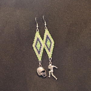 May include: A pair of dangle earrings featuring diamond-shaped beaded patterns in lime green, dark blue, and white. One earring has a silver-toned football helmet charm, while the other features a silver-toned football player figure charm.