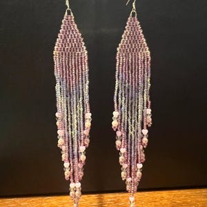 May include: A pair of long, beaded earrings in shades of purple, lavender, and white. The earrings have a fringe design with small beads and silver-colored hooks. The earrings are displayed against a dark background.