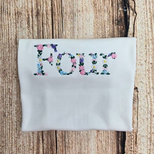 May include: White t-shirt with the word "FOUR" embroidered in pink, blue and purple flowers.