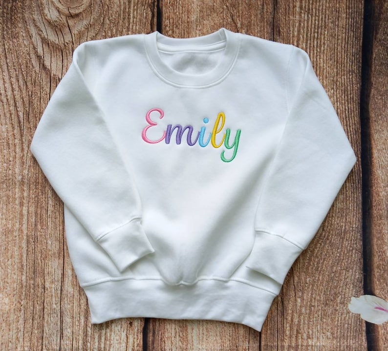 May include: White sweatshirt with the name "Emily" embroidered in rainbow colours.