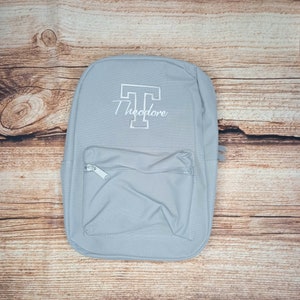 Peut inclure: Un sac à dos en toile gris avec une poche zippée à l'avant. Le sac à dos a une lettre "T" brodée en blanc et le nom "Theodore" sur le devant.