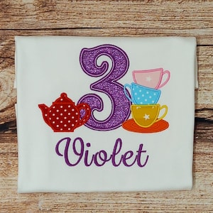 May include: Purple glittery number three with a red teapot and a stack of colourful teacups with a purple name "Violet" on a white shirt.