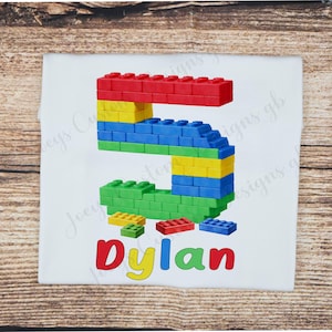 May include: White t-shirt featuring a colorful number 5 constructed from toy building blocks. The name "Dylan" is written in red, yellow, and green below the number. Additional blocks are scattered around the number.