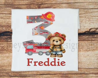 May include: A white t-shirt with a red fire truck, a firefighter teddy bear, and the number 3. The number 3 is made of a gray pattern of fire trucks. The shirt is personalized with the name "Freddie".