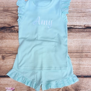 May include: A light blue pyjama set with a ruffled trim. The top has short sleeves and the bottom is shorts. The top has the name "Amy" embroidered on the front.