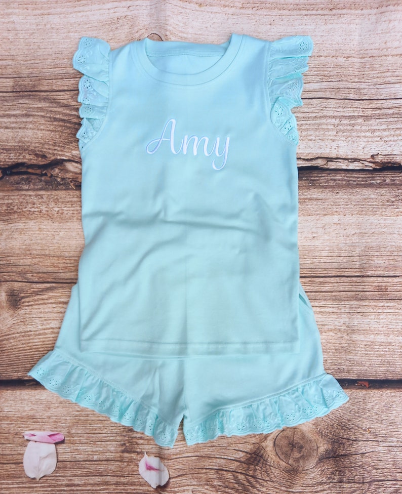 May include: A light blue pyjama set with a ruffled trim. The top has short sleeves and the bottom is shorts. The top has the name "Amy" embroidered on the front.