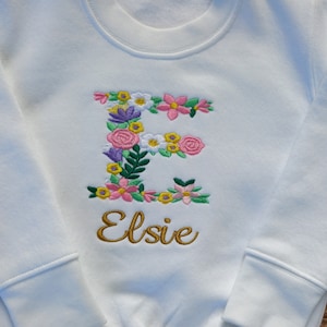 May include: White sweatshirt with a floral embroidered letter E and the name Elsie in gold embroidery.