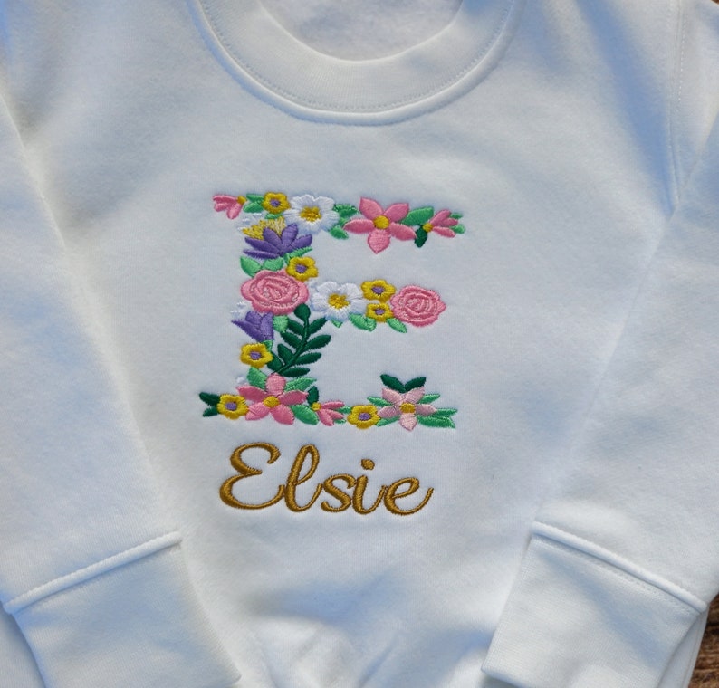 May include: White sweatshirt with a floral embroidered letter E and the name Elsie in gold embroidery.