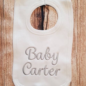 Puede incluir: Babero blanco para bebé con una abertura para el cuello y el texto bordado "Baby Carter" en escritura gris. El babero está hecho de tela suave y tiene un borde inferior redondeado.