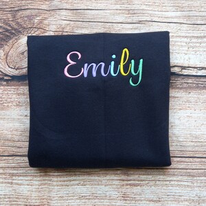 May include: Black folded fabric with the name "Emily" embroidered in rainbow colours.