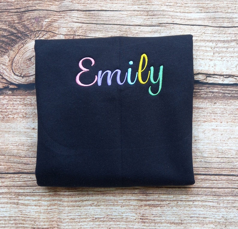 May include: Black folded fabric with the name "Emily" embroidered in rainbow colours.
