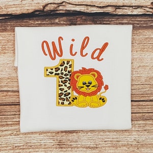 Puede incluir: Un body blanco con el texto "Wild 1" en naranja. El número 1 está decorado con un estampado de leopardo. Un león de dibujos animados con un cuerpo amarillo, una melena naranja y una cola negra está sentado junto al número.