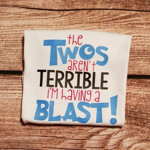 Puede incluir: Camiseta blanca con el texto "the Twos aren't TERRIBLE i'm having a BLAST!" en letras azules, negras y rosas.