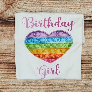 May include: White t-shirt with the text "Birthday Girl" in pink lettering. A rainbow heart-shaped pop-it toy is centered on the shirt.