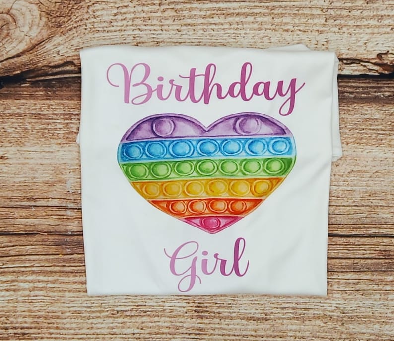 May include: White t-shirt with the text "Birthday Girl" in pink lettering. A rainbow heart-shaped pop-it toy is centered on the shirt.