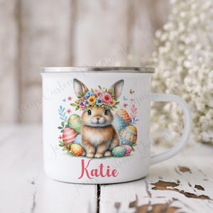 May include: White enamel mug featuring a colourful Easter bunny wearing a flower crown and surrounded by Easter eggs. The mug has the name "Katie" printed on the bottom.
