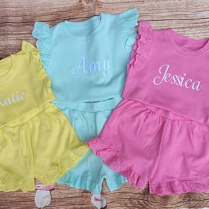 May include: Three sets of personalised baby girl pyjamas with ruffled sleeves and shorts. The pyjamas are yellow, light blue, and pink with white embroidered names "Katlie", "Amy", and "Jessica".