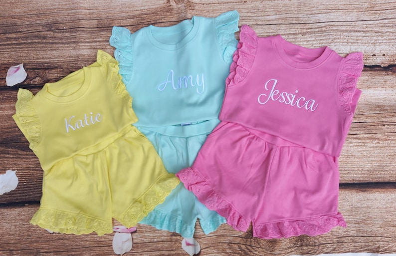 May include: Three sets of personalised baby girl pyjamas with ruffled sleeves and shorts. The pyjamas are yellow, light blue, and pink with white embroidered names "Katlie", "Amy", and "Jessica".